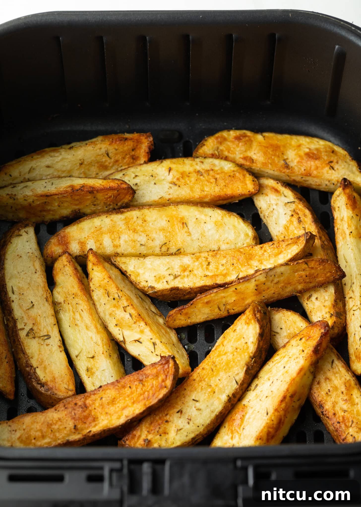A close-up view of beautifully crispy, golden-brown air-fried potato wedges nestled in a black air fryer basket, steam gently rising.