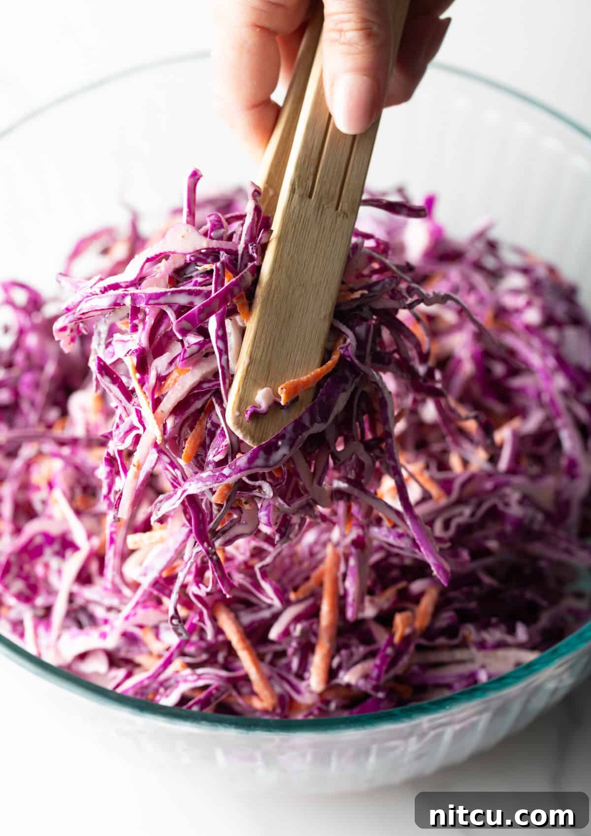 Red cabbage slaw is being tossed with wooden tongs in a glass bowl.