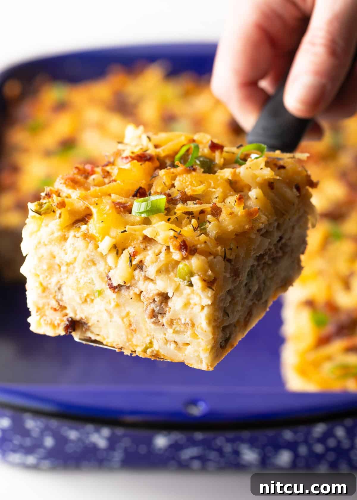 A slice of hashbrown breakfast casserole is being held by a metal spatula above a blue baking dish, showcasing its cheesy layers and golden-brown top.