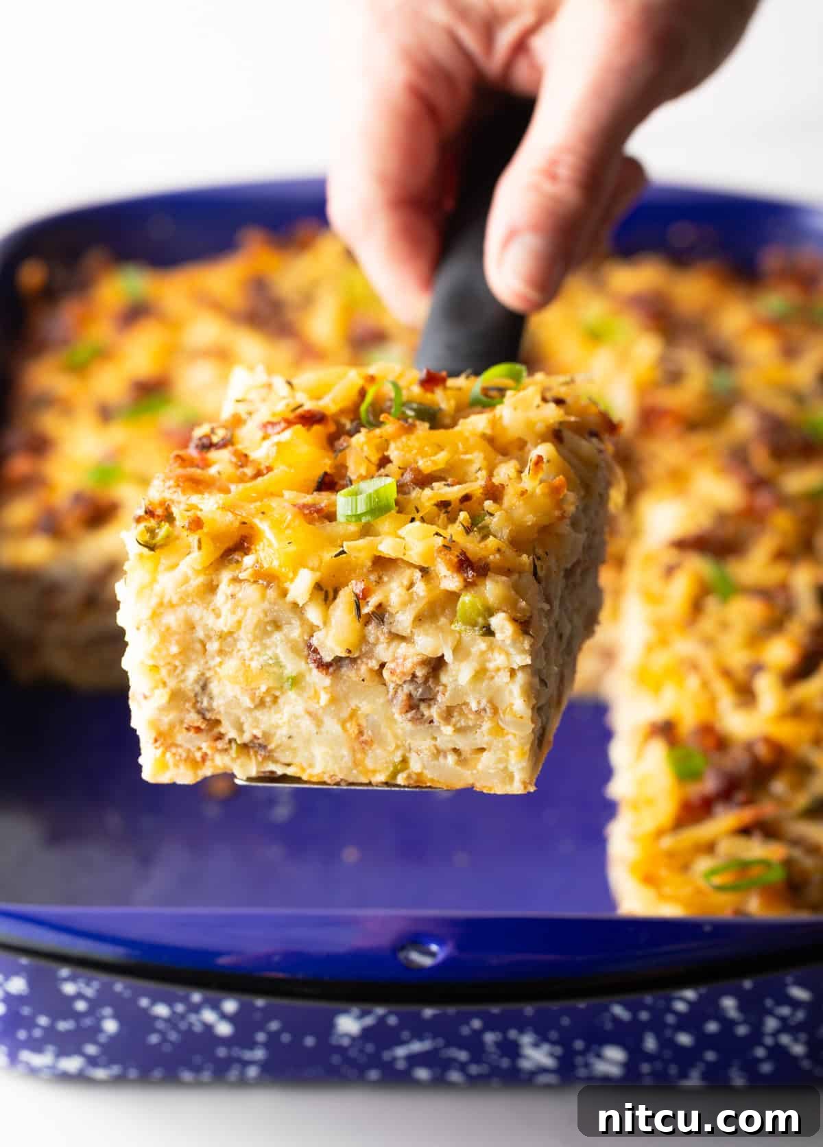 A hand holds up a perfectly square slice of golden-brown breakfast casserole towards the camera, highlighting its delicious texture.