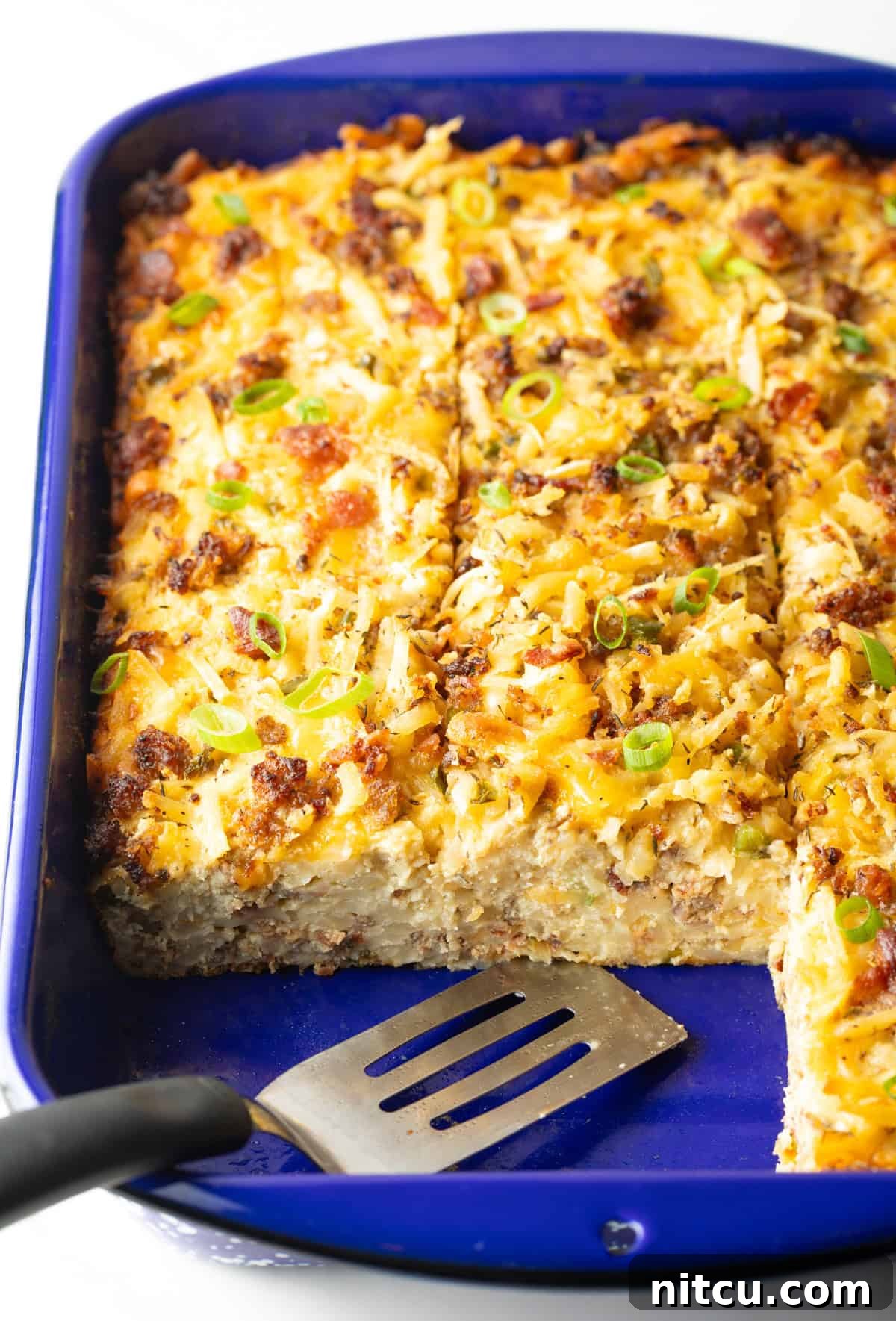 A freshly baked breakfast hash brown casserole, with two slices already served, is perfectly presented in a blue baking dish alongside a metal spatula.