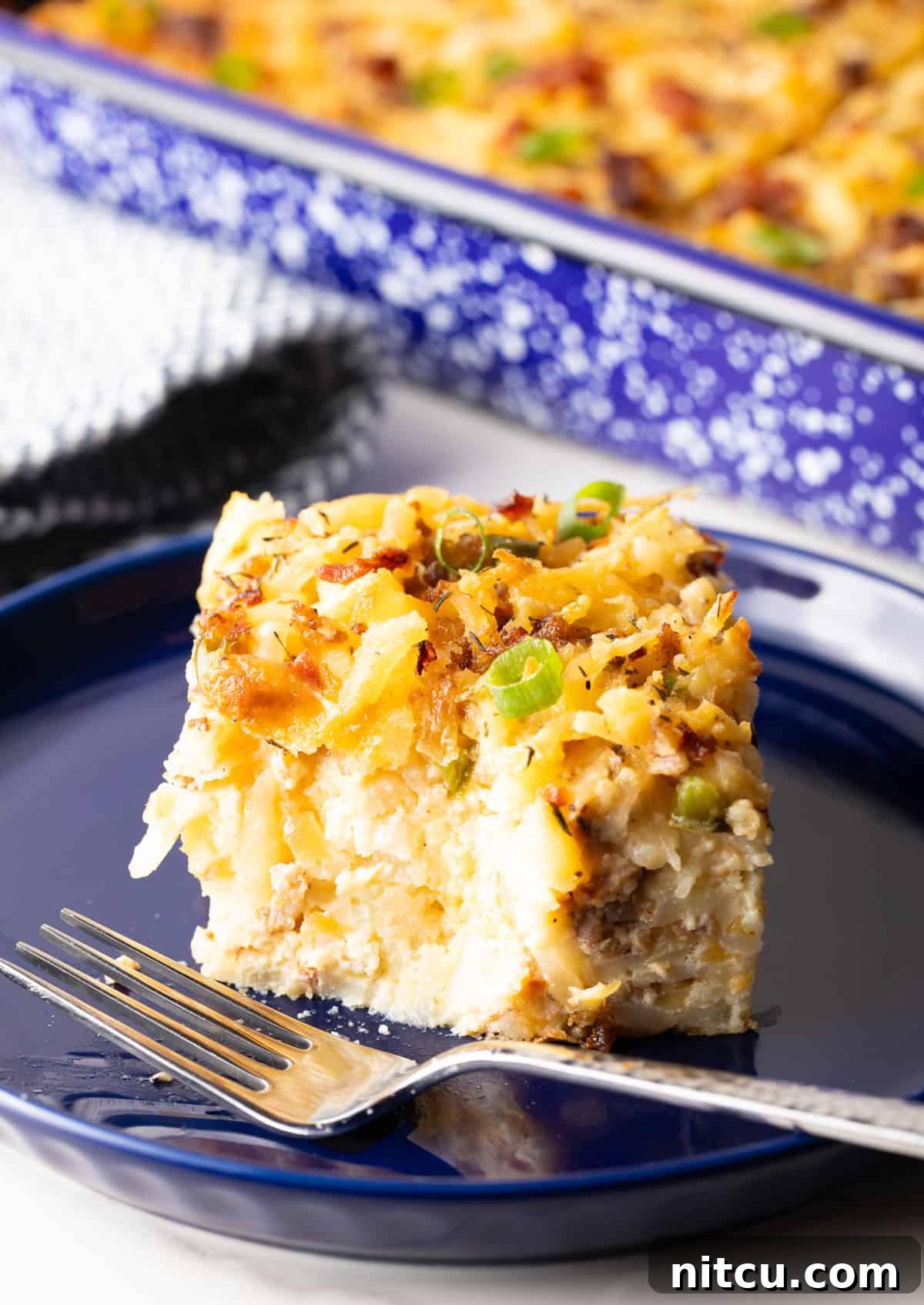 A delectable slice of breakfast casserole with hash browns rests on a blue plate, with a fork poised to take another tempting bite.