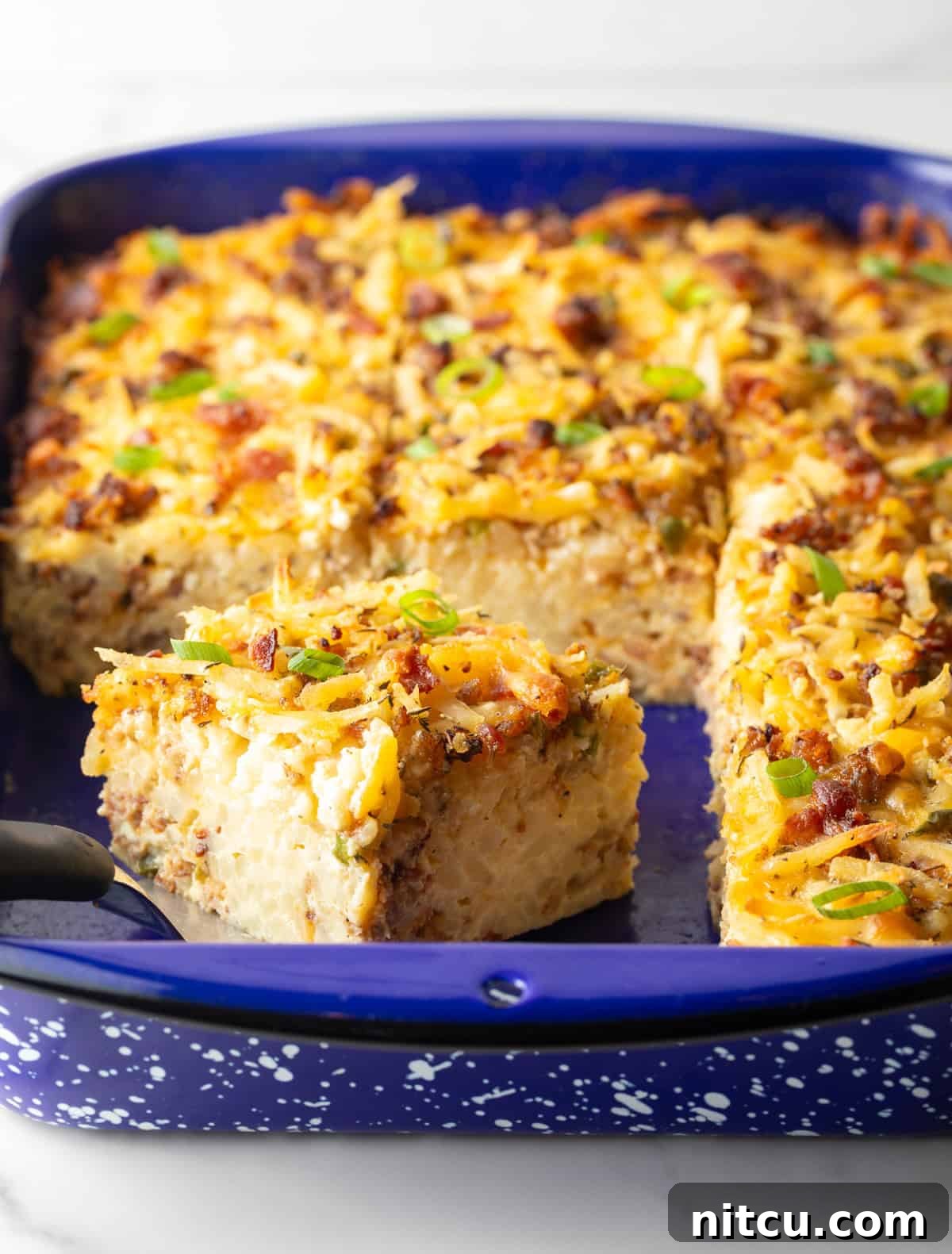 A full hash brown breakfast casserole in a vibrant blue baking dish, with a metal spatula lifting a single perfect slice.
