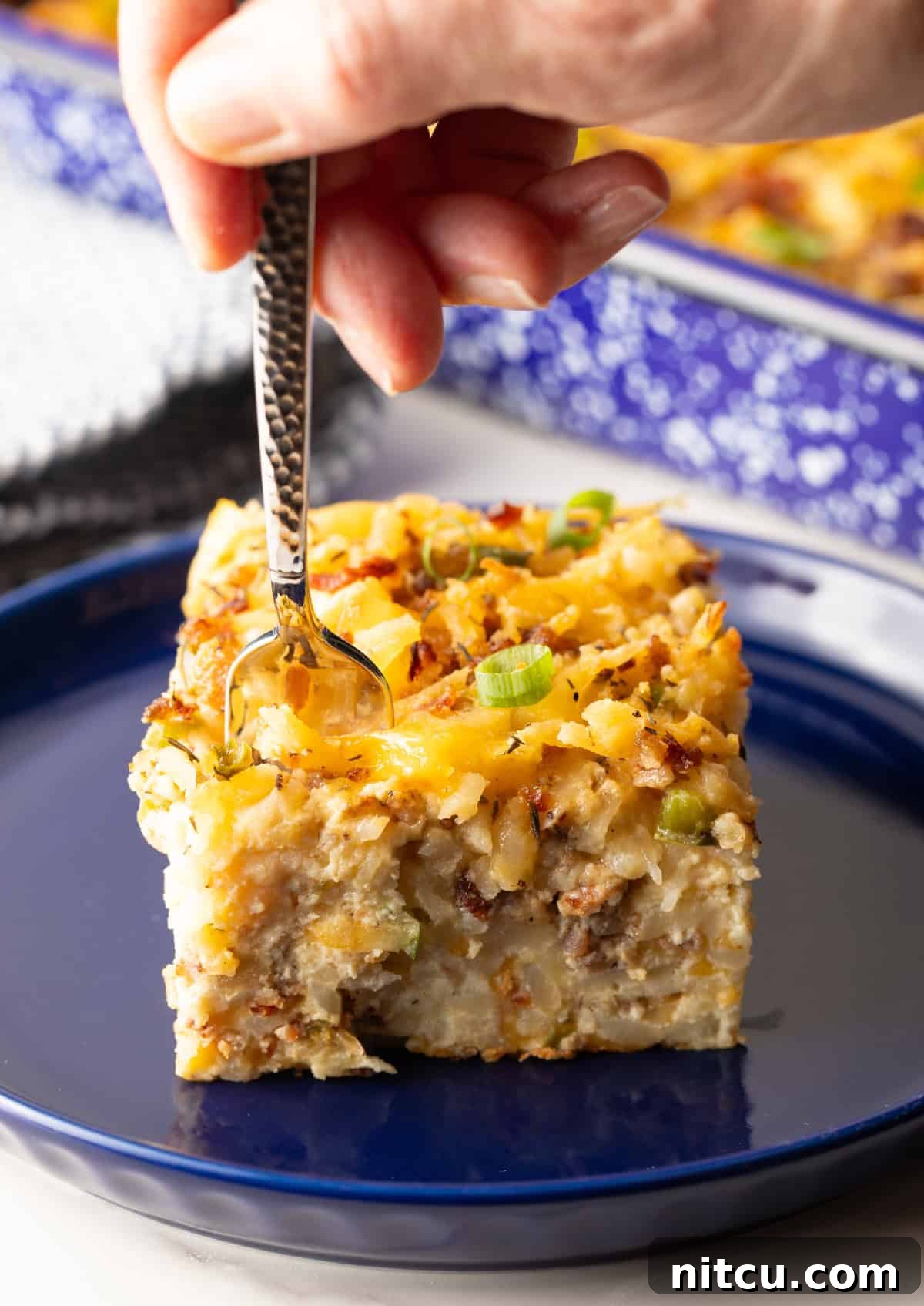 A delicious slice of hash brown breakfast casserole on a blue plate, with a fork taking a mouth-watering bite out of it.
