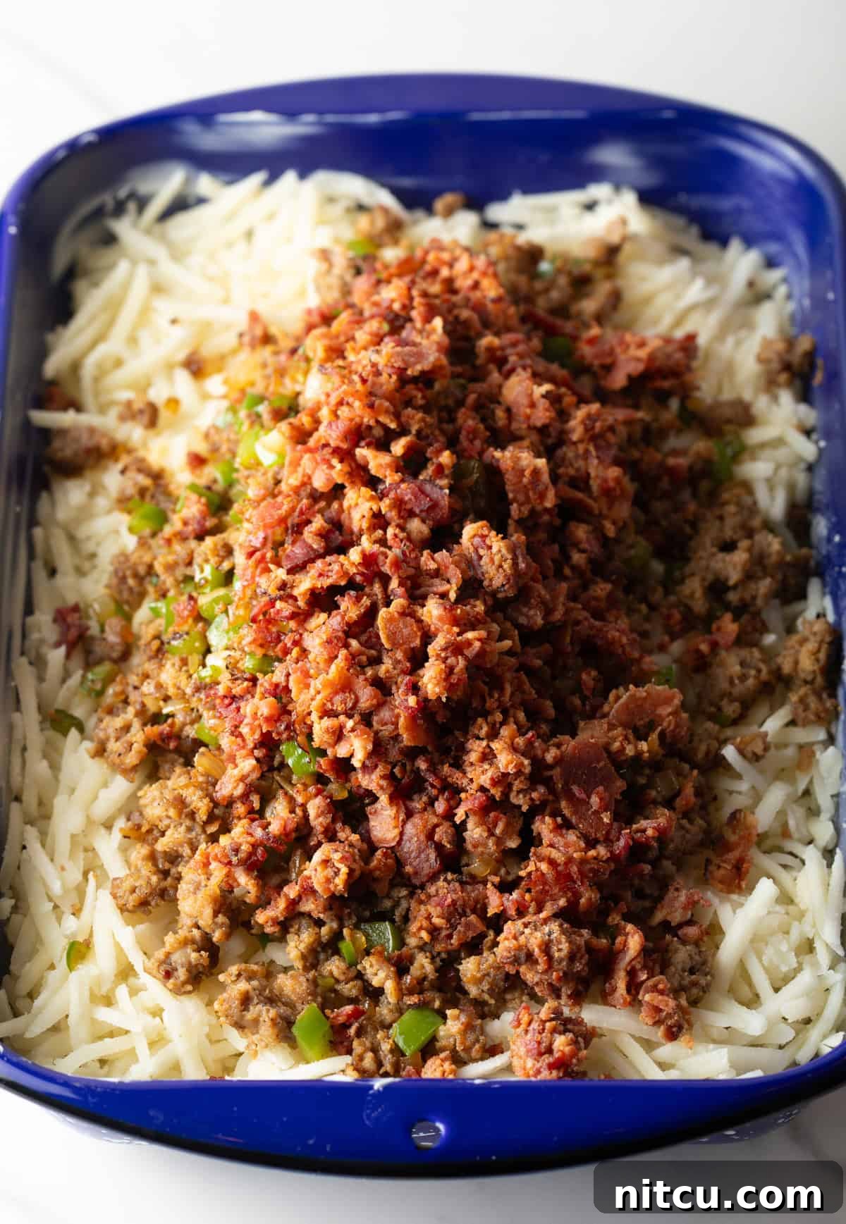 Frozen hash browns form the base layer, topped with a delicious mixture of cooked sausage and bacon, ready for the next step in a blue casserole dish.