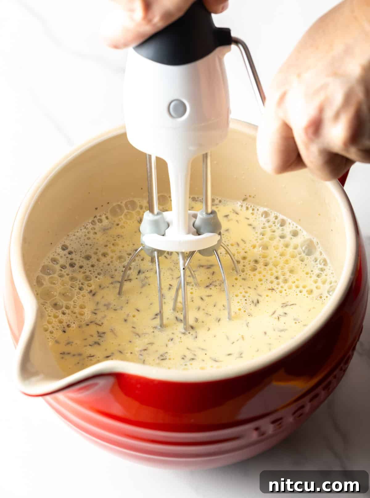 A whisk vigorously mixes an egg and cream mixture in a vibrant red bowl, preparing it for the casserole.