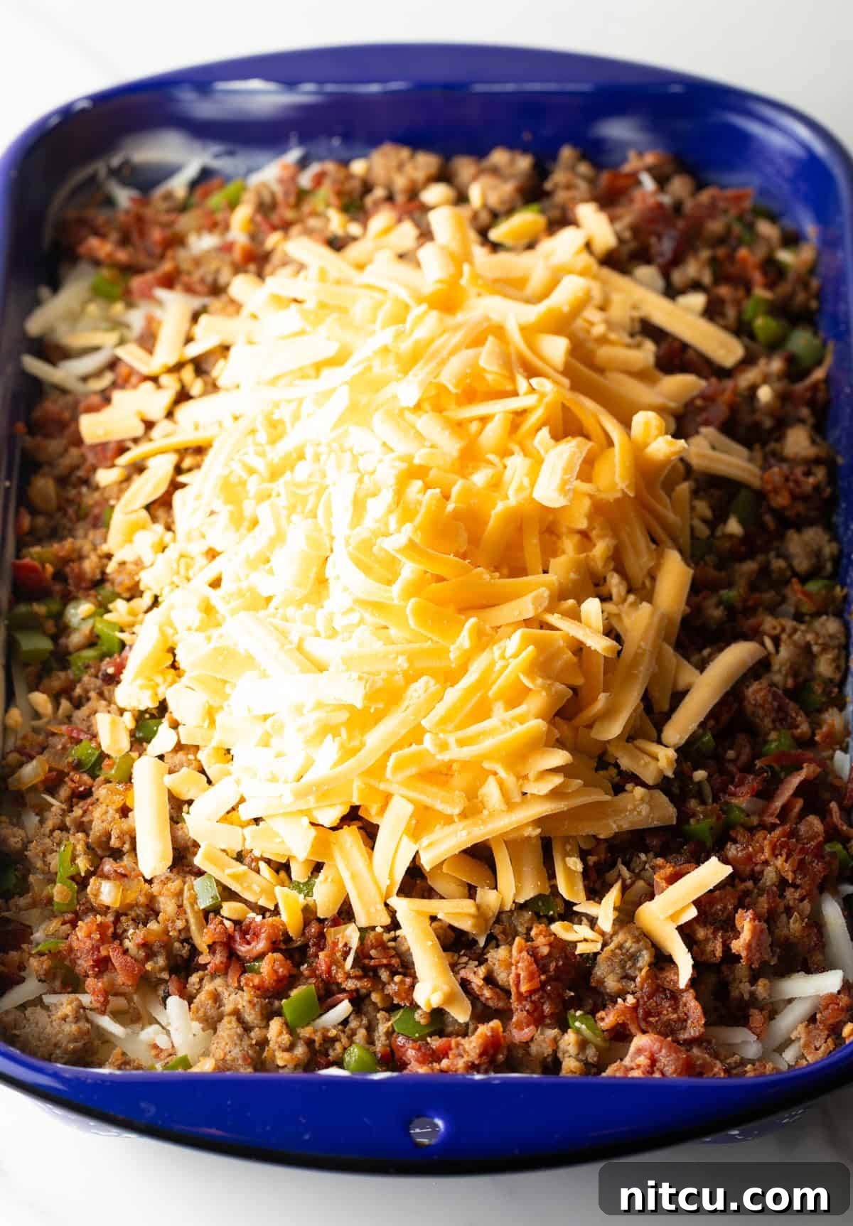 Shredded cheddar cheese is being added generously to a casserole dish already filled with layers of hash browns and seasoned sausage.