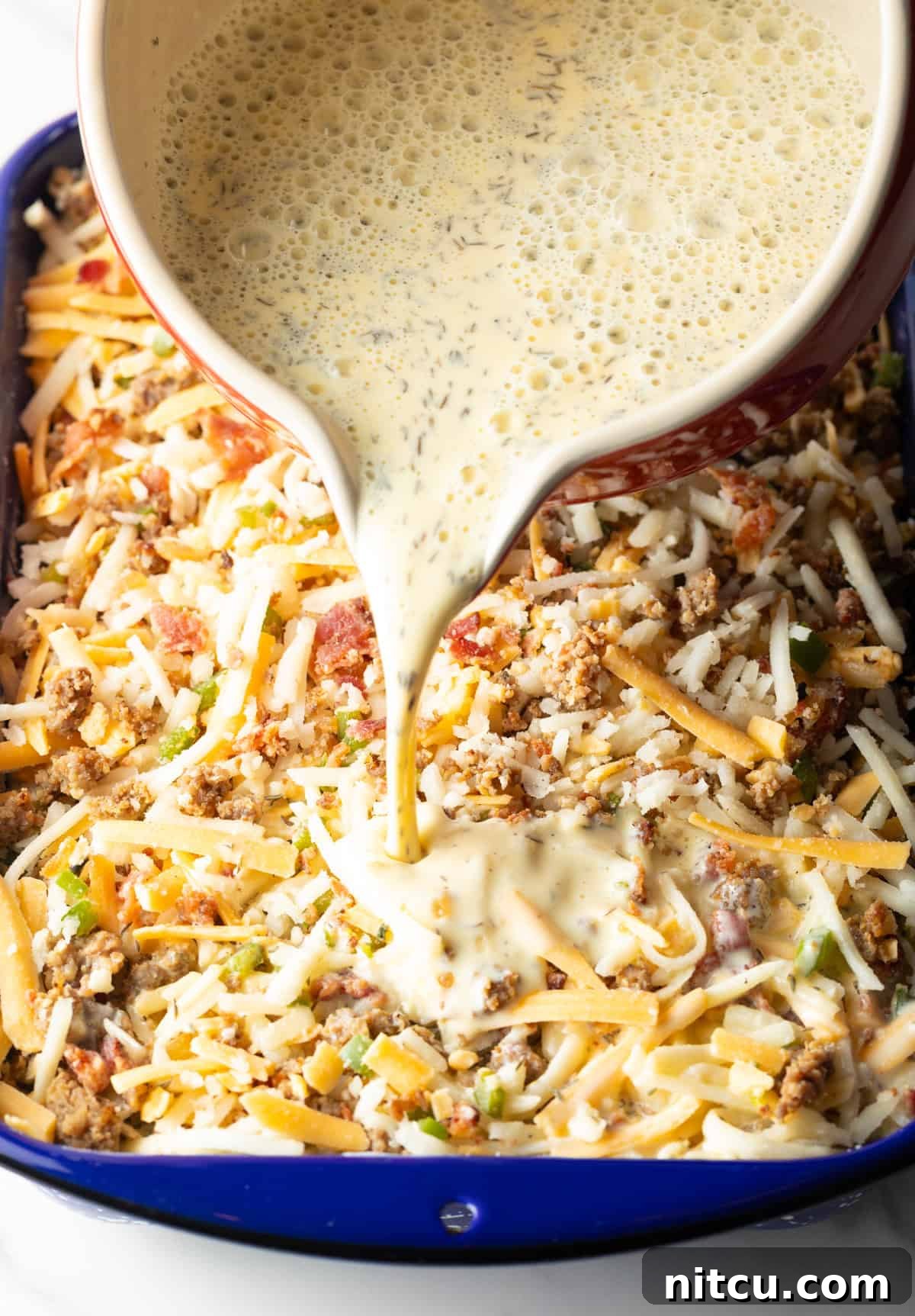 A smooth egg and cream mixture is gracefully poured over the assembled ingredients for a hash brown breakfast casserole, ensuring every component is saturated.
