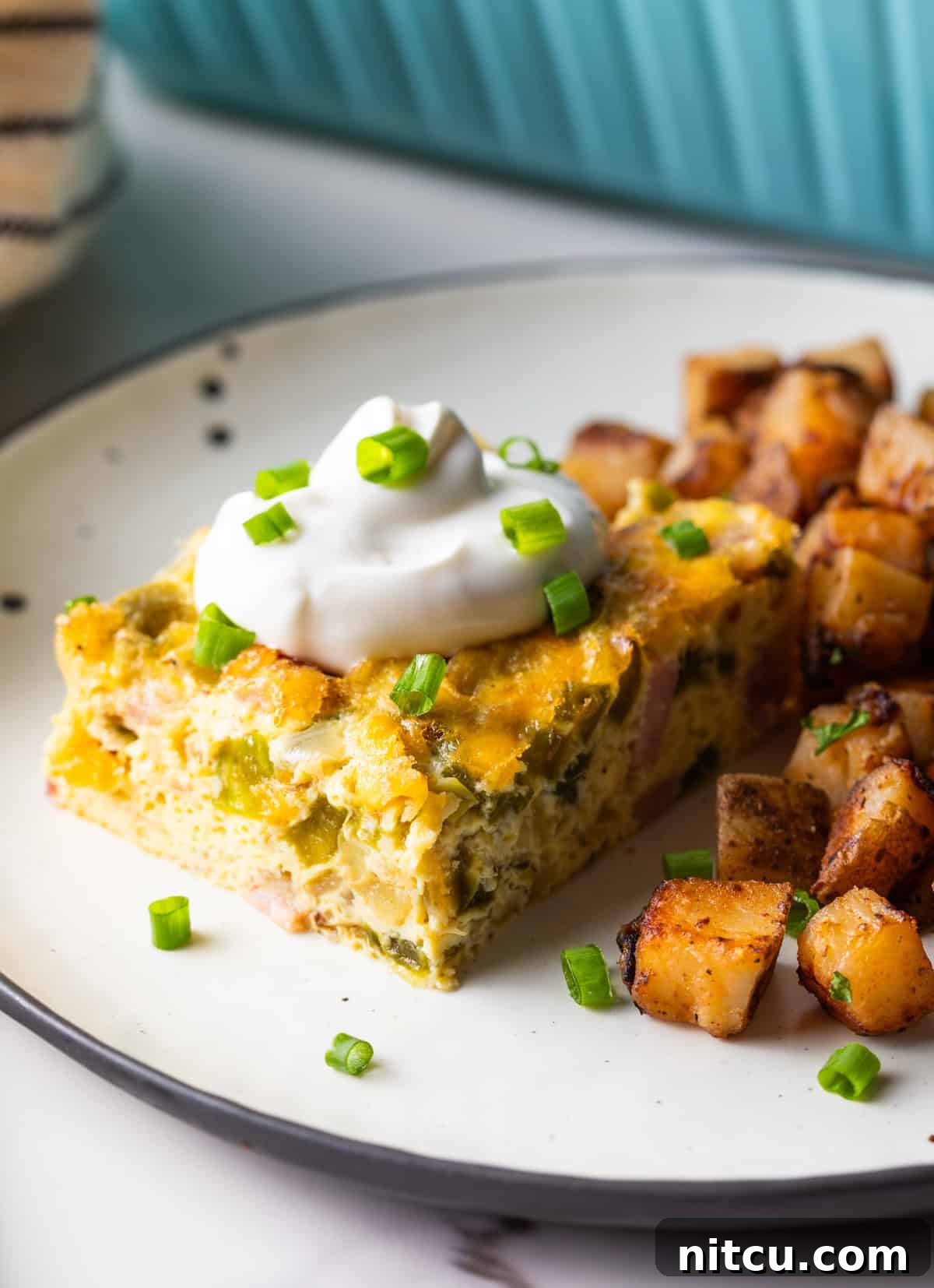 A slice of baked Denver omelet casserole topped with sour cream, next to a serving of home fries on a white plate.