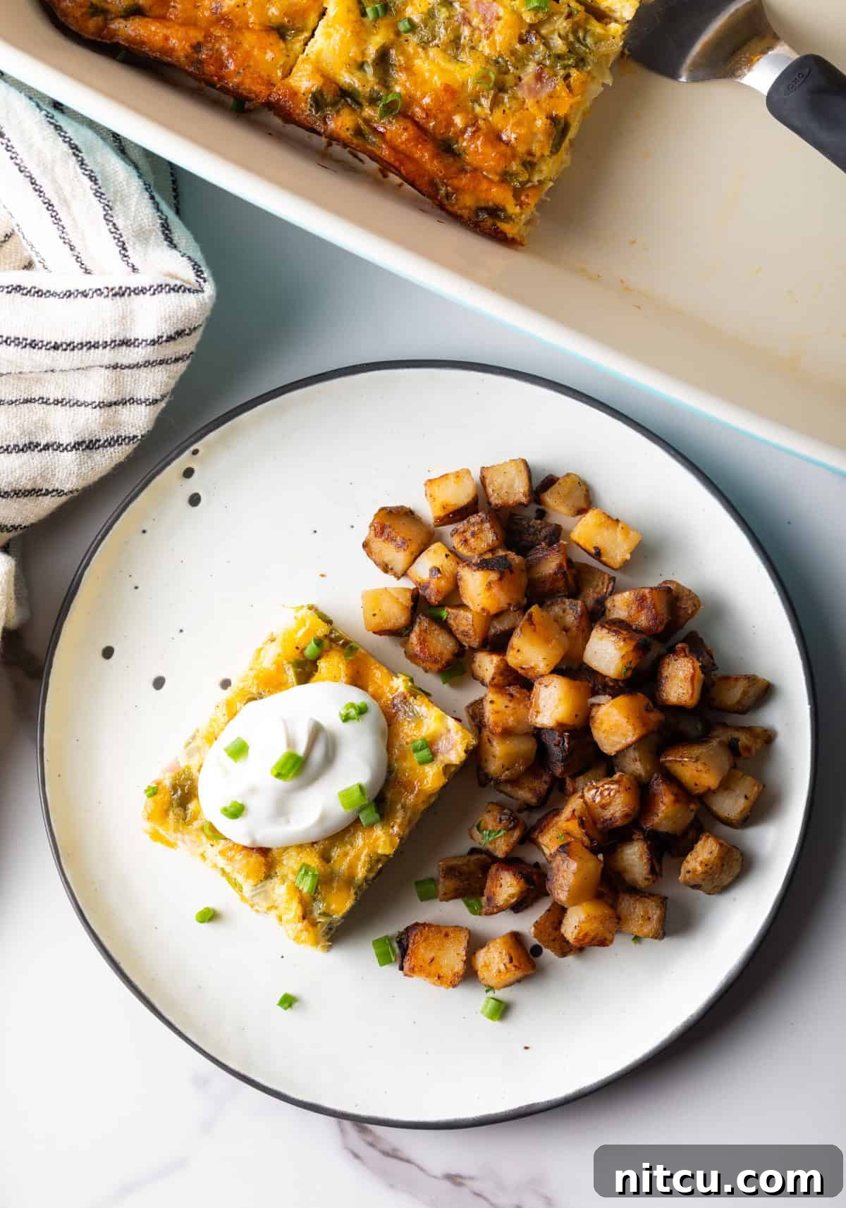 An overhead shot of a slice of baked Denver omelet casserole topped with sour cream, next to a serving of home fries on a white plate.