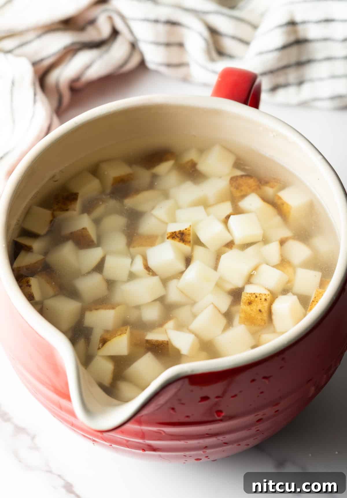Cubed potatoes sitting in an ice and water bath in a red measuring bowl.