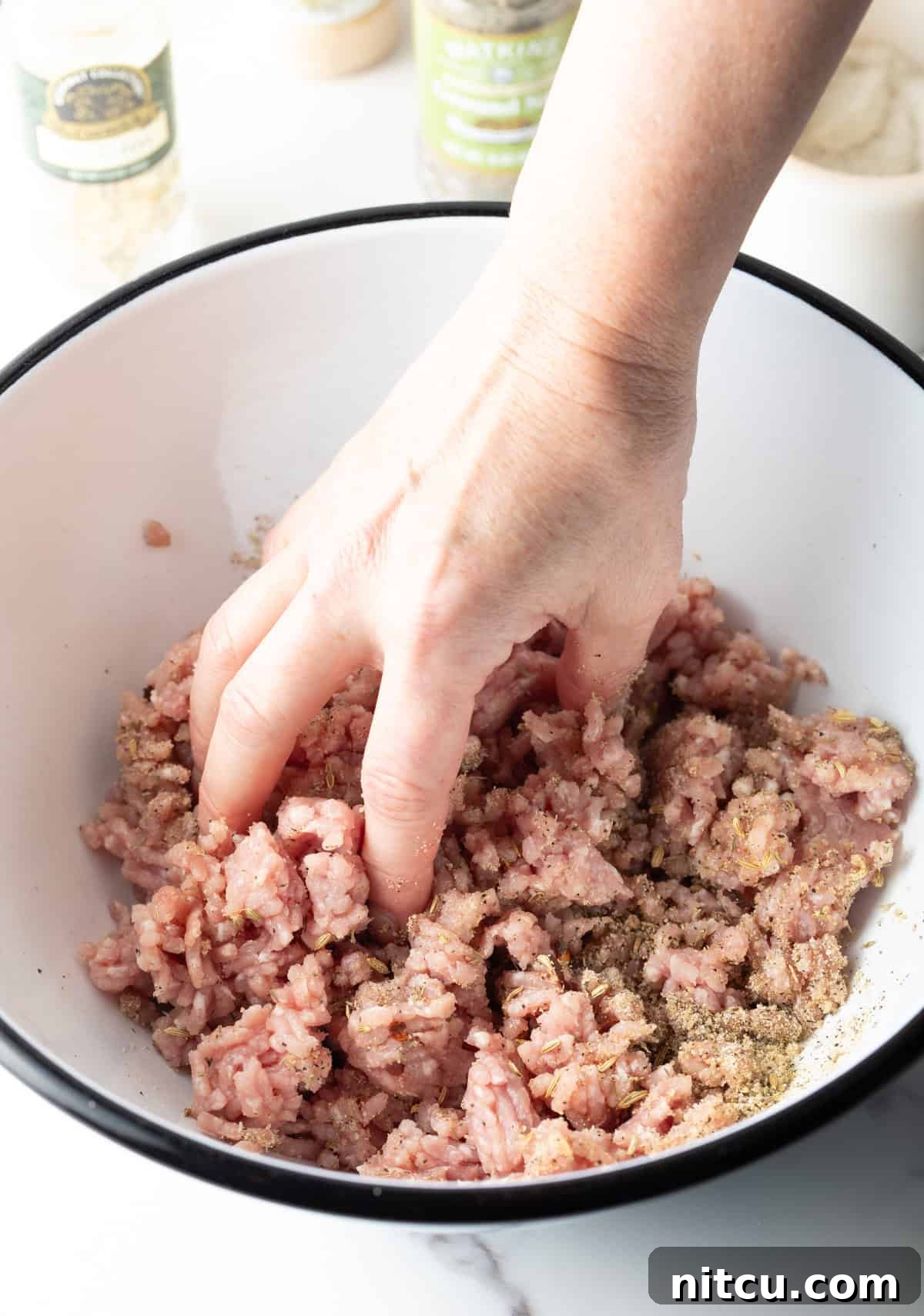 A hand is mixing a ground pork and seasoning mixture together in a white bowl.