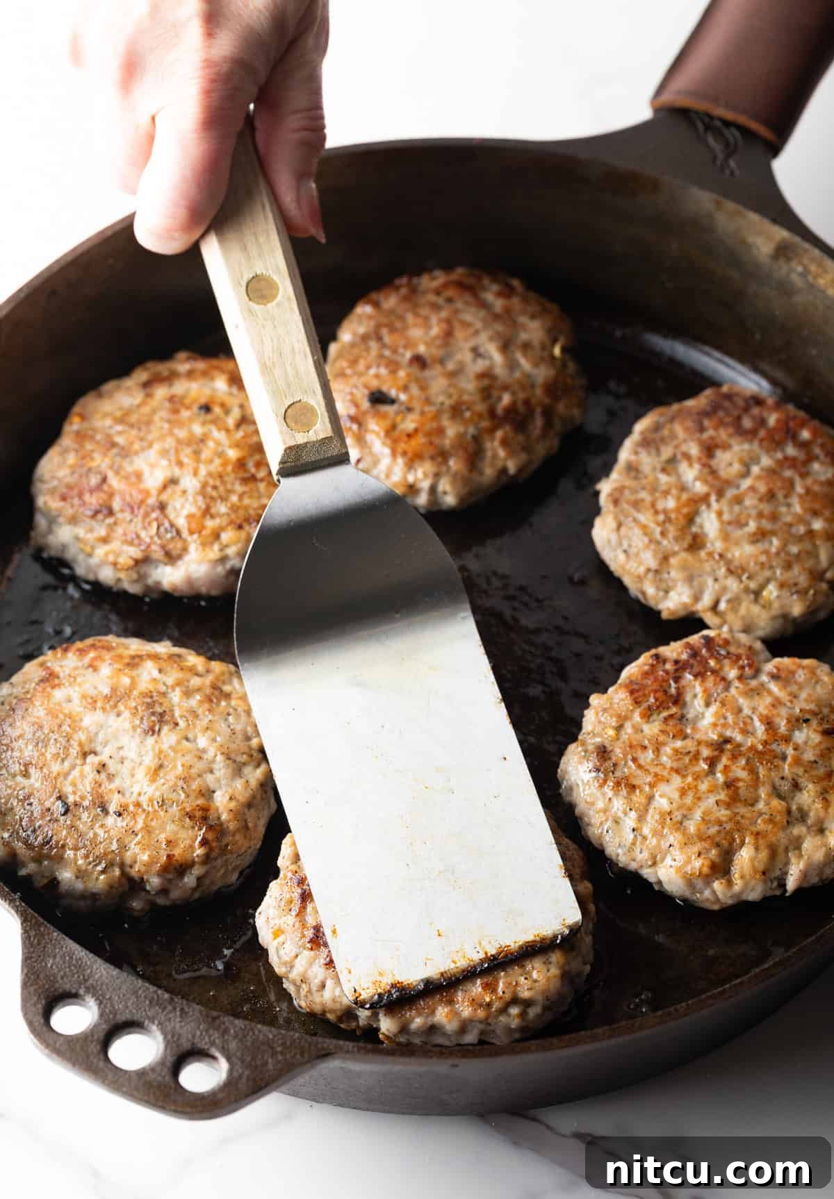 Homemade breakfast sausage patties in a black skillet, with a metal spatula pressing one of the patties.