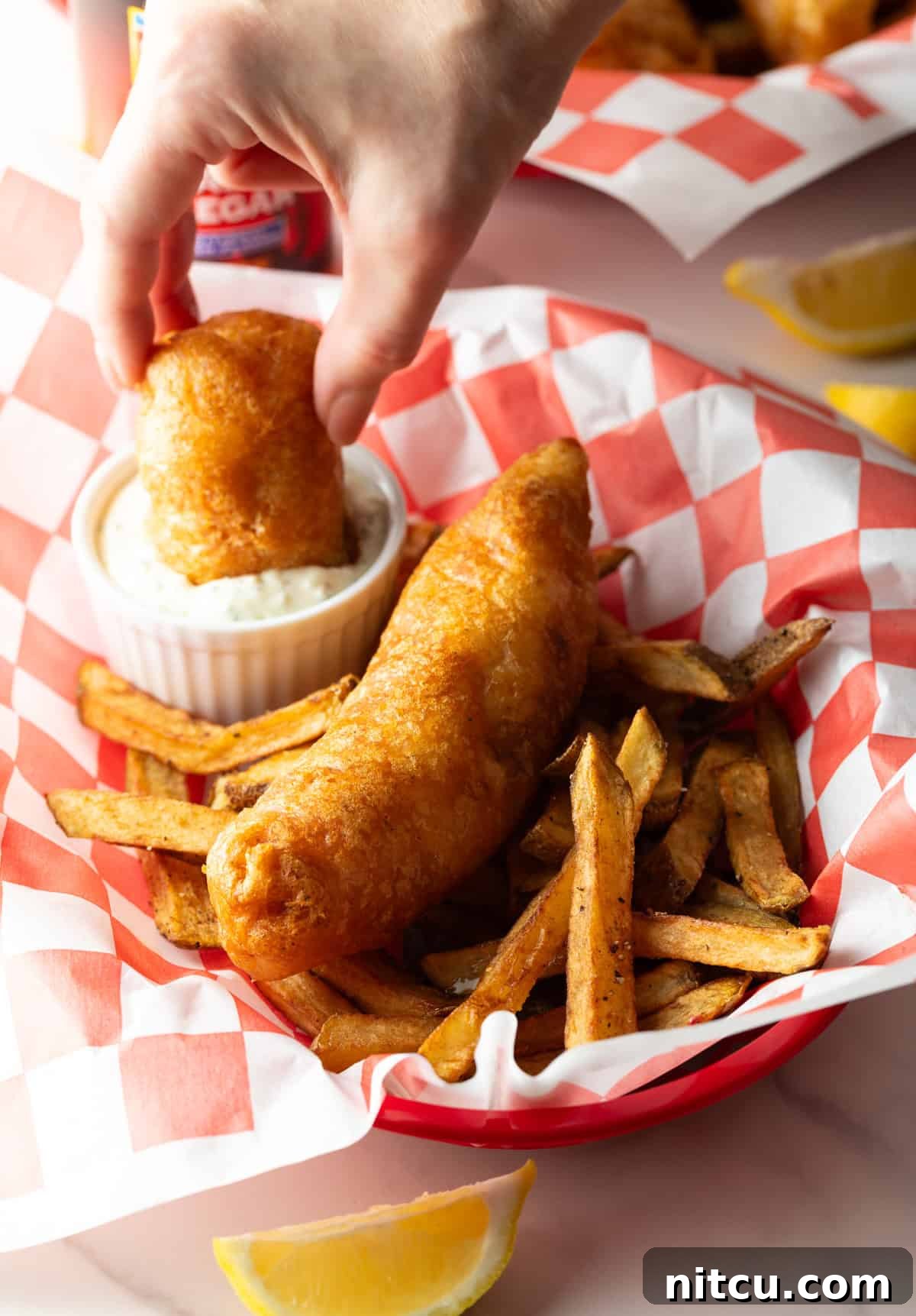 A hand is dunking a piece of crispy fried fish into a ramekin filled with sauce.