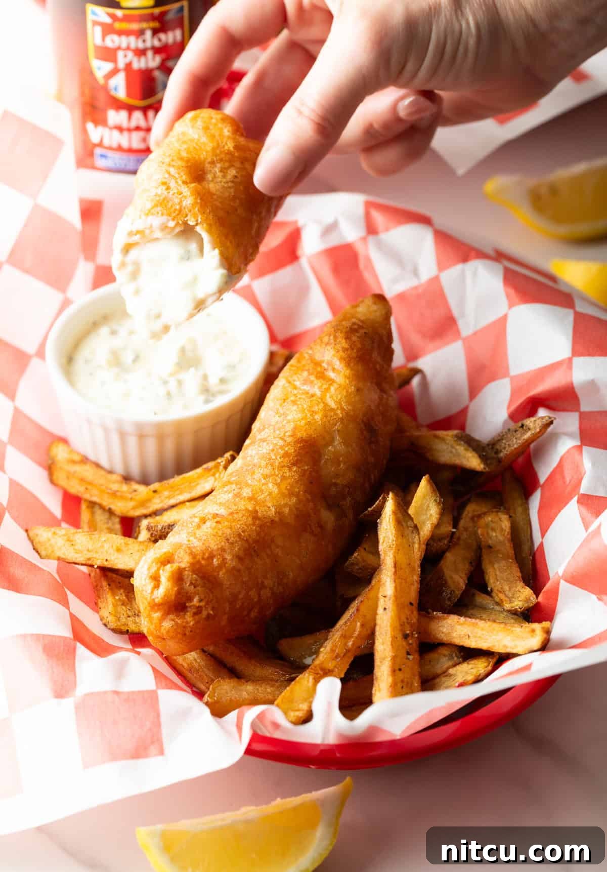 A fish and chip recipe sitting in a paper lined basket, with a piece of fish being dunked into a ramekin of sauce.