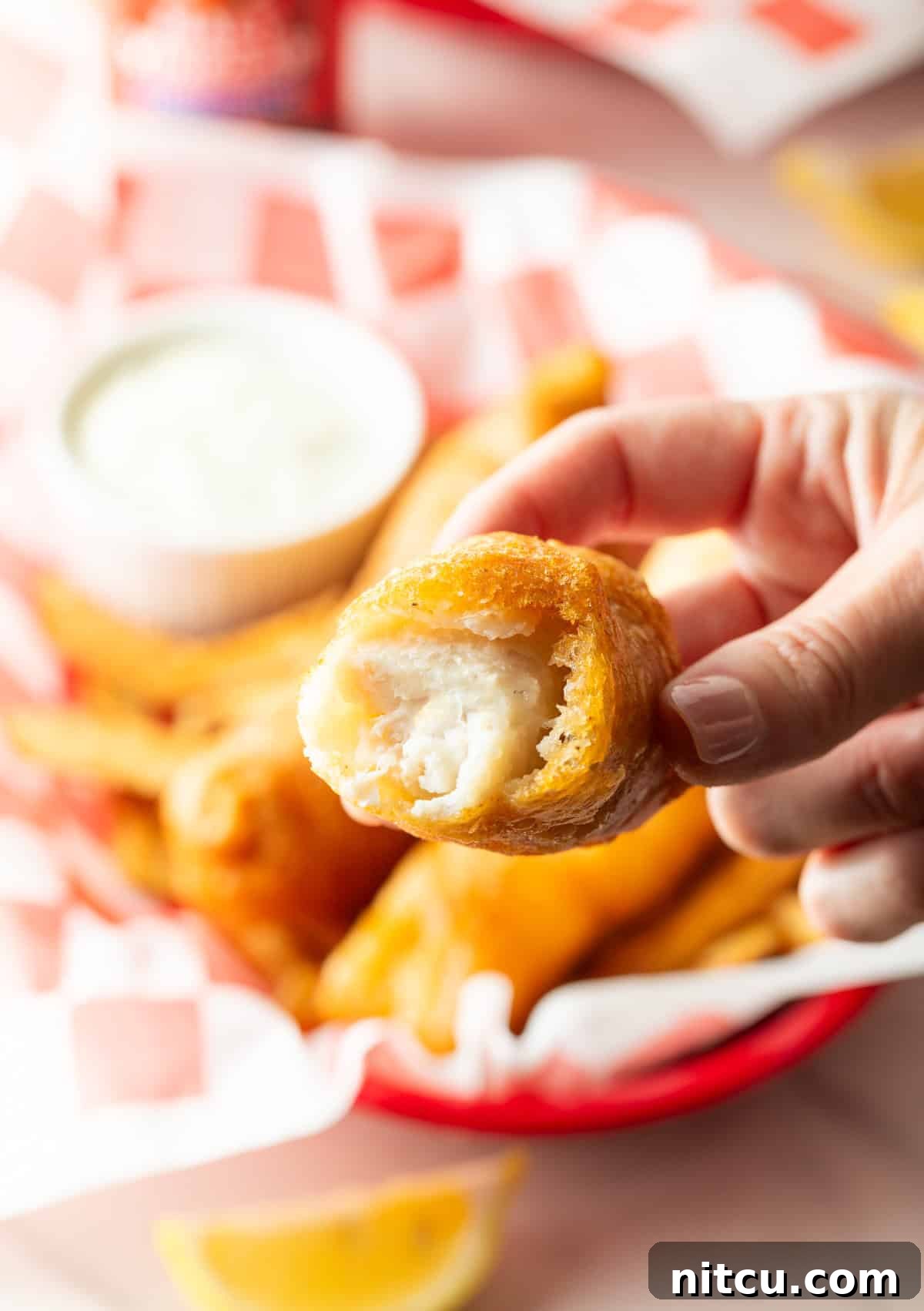 A hand is holding a piece of battered fried fish with a bite taken out, and it is being held over a basket of fish and chips.