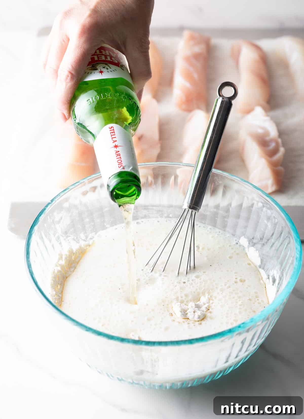 A bottle of beer being poured into a fried fish batter in a glass bowl.