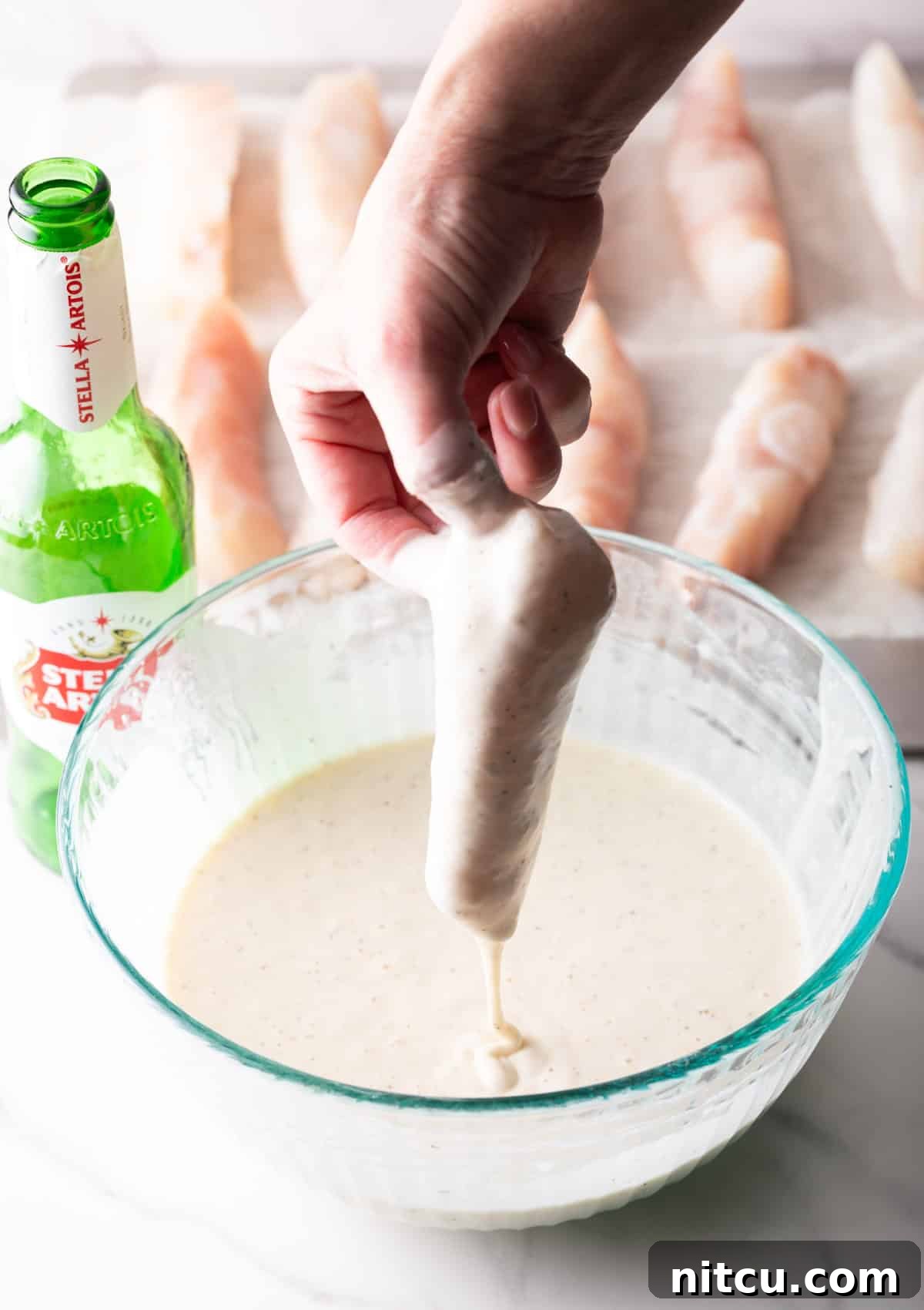 A hand is holding a fish fillet that is being dipped in a fish and chips batter.
