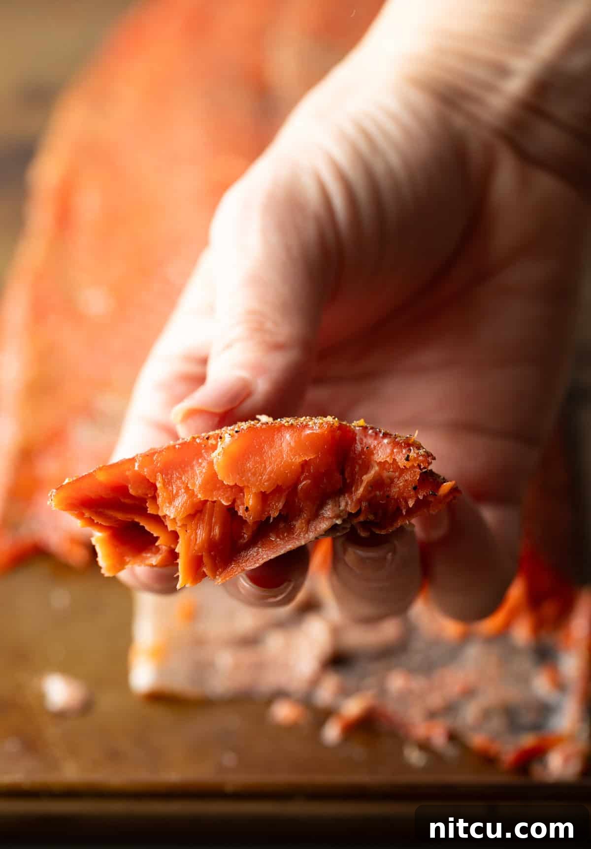 A hand holding a piece of smoked salmon, above the full fillet of cold smoked salmon.