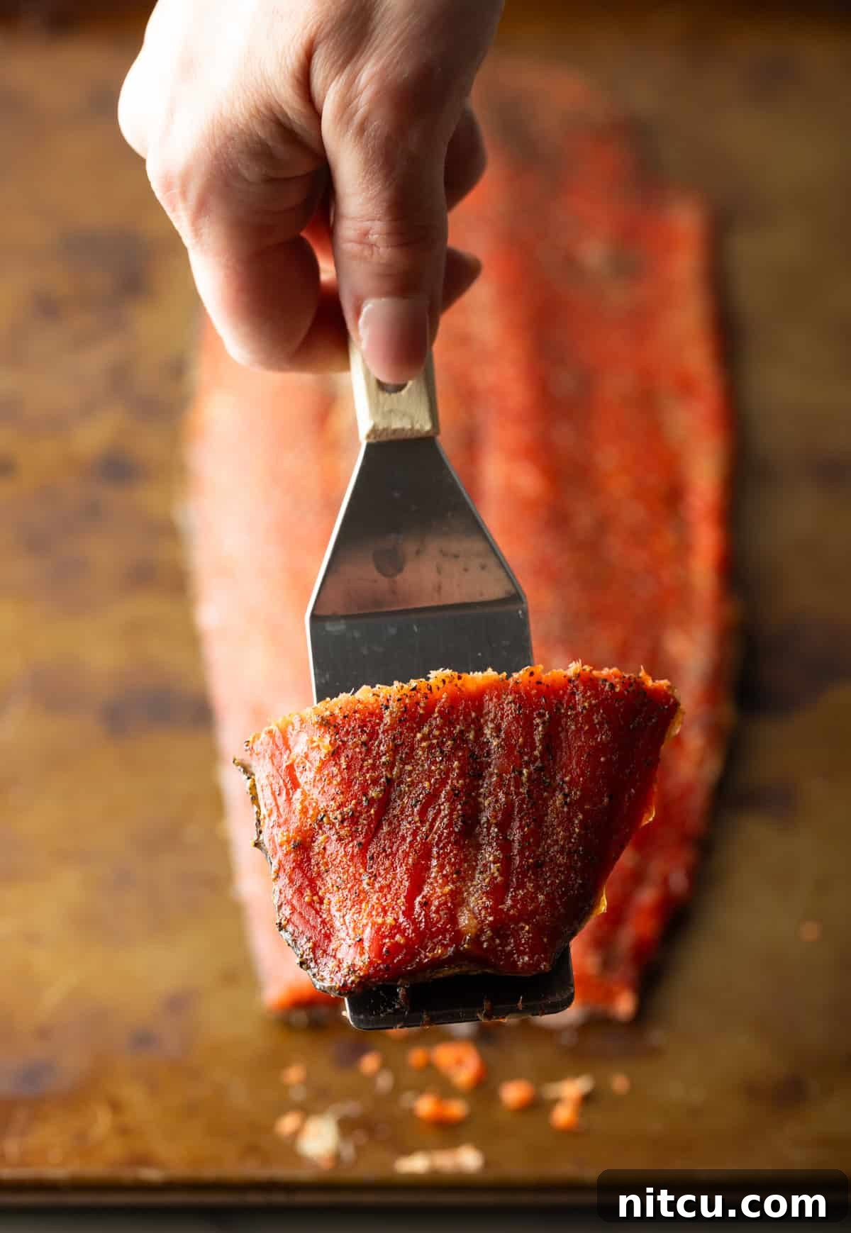 A metal spatula is holding a slice of easy cold smoked salmon above the full fillet of salmon.