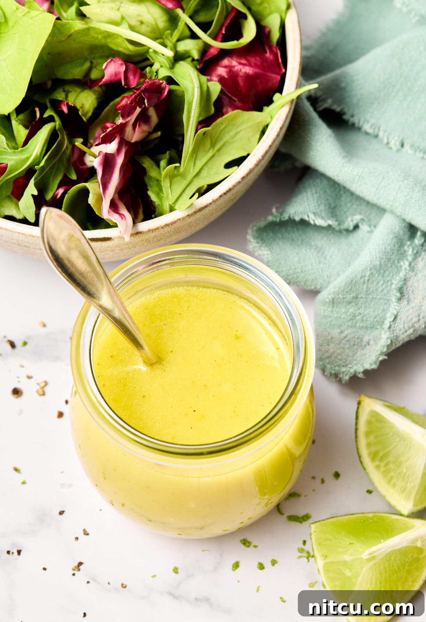 An overhead shot of a lime vinaigrette recipe in a glass jar, next to wedges of lime and a salad.