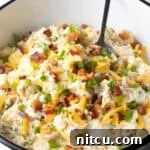 A loaded baked potato salad recipe topped with bacon, cheese, and chives, sitting in white bowl with a spoon.