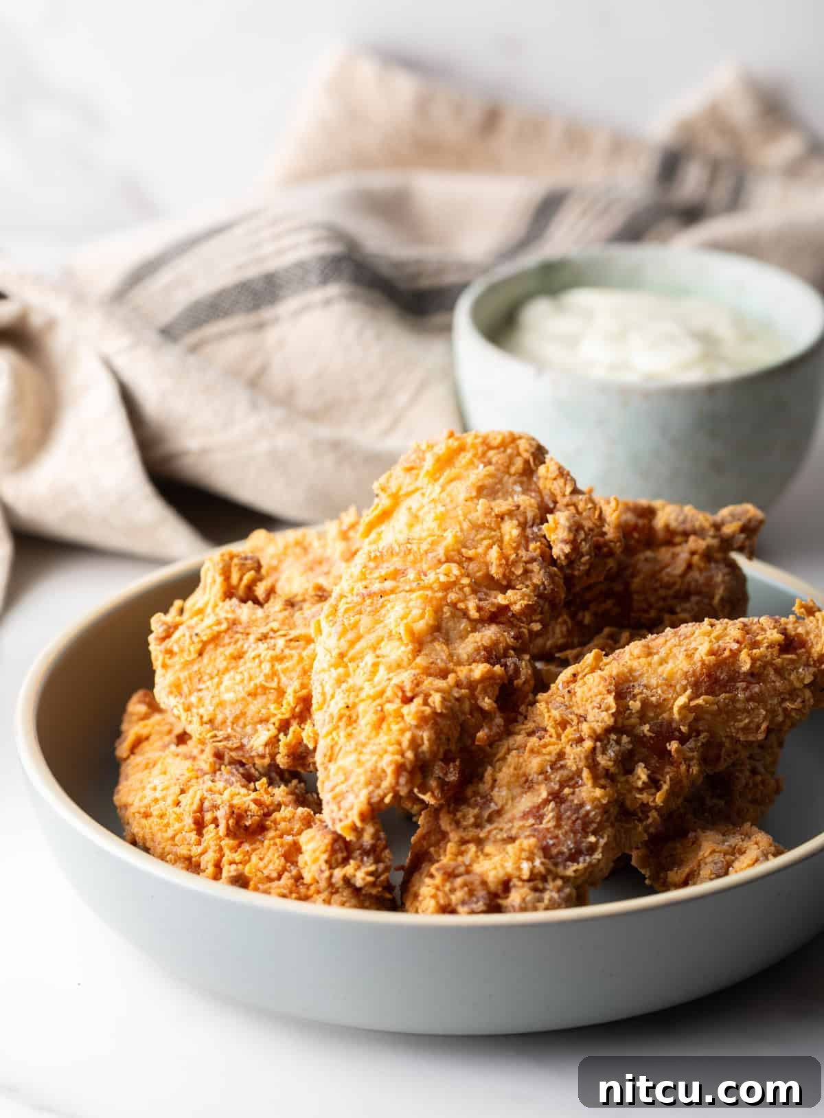 Crispy Deep-Fried Chicken Tenders Recipe That Delivers 10 A white bowl filled with deep fried chicken tenders, with a ramekin of white sauce on the side.