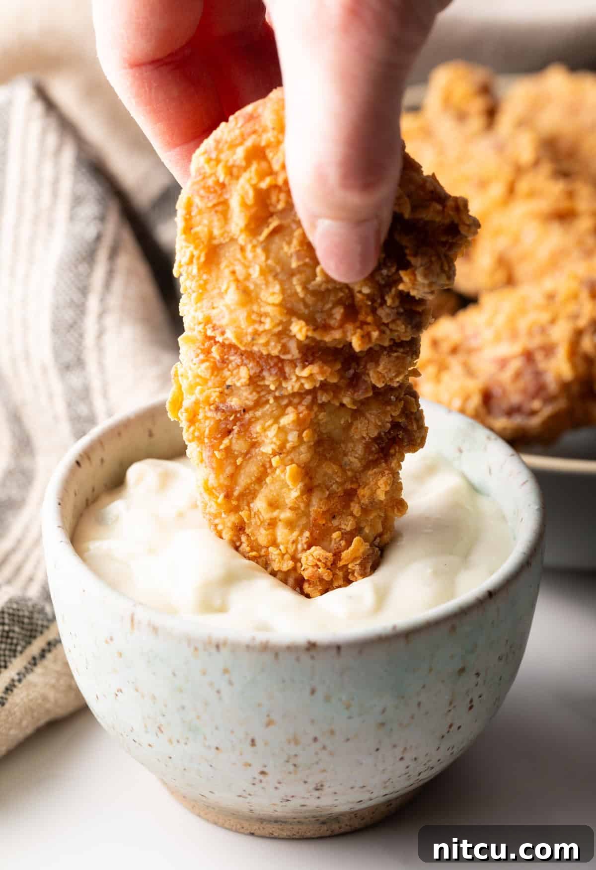 Crispy Deep-Fried Chicken Tenders Recipe That Delivers 3 A homemade fried chicken tender is being dipped into a ramekin of white sauce.