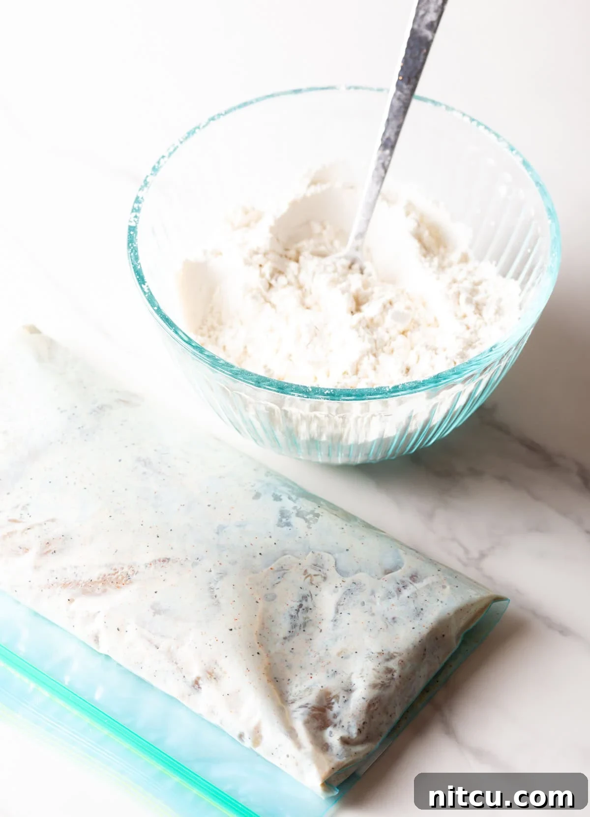 Crispy Deep-Fried Chicken Tenders Recipe That Delivers 6 A glass bowl filled with a flour mixture sitting next to a bag of raw chicken tenders marinating in a buttermilk mixture.