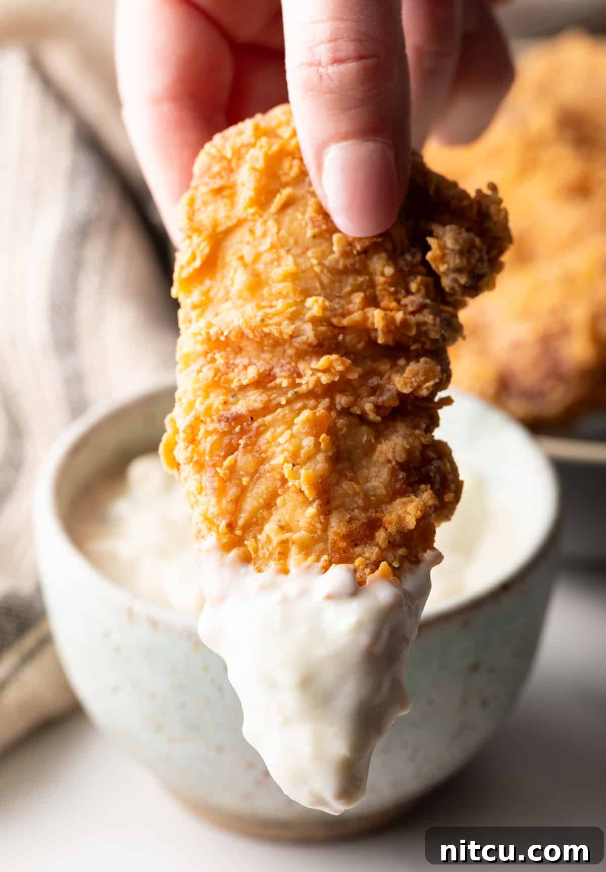 Crispy Deep-Fried Chicken Tenders Recipe That Delivers 8 A homemade deep fried chicken tender is being dipped into a ramekin of white sauce, with the sauce dripping over the side of the ramekin.