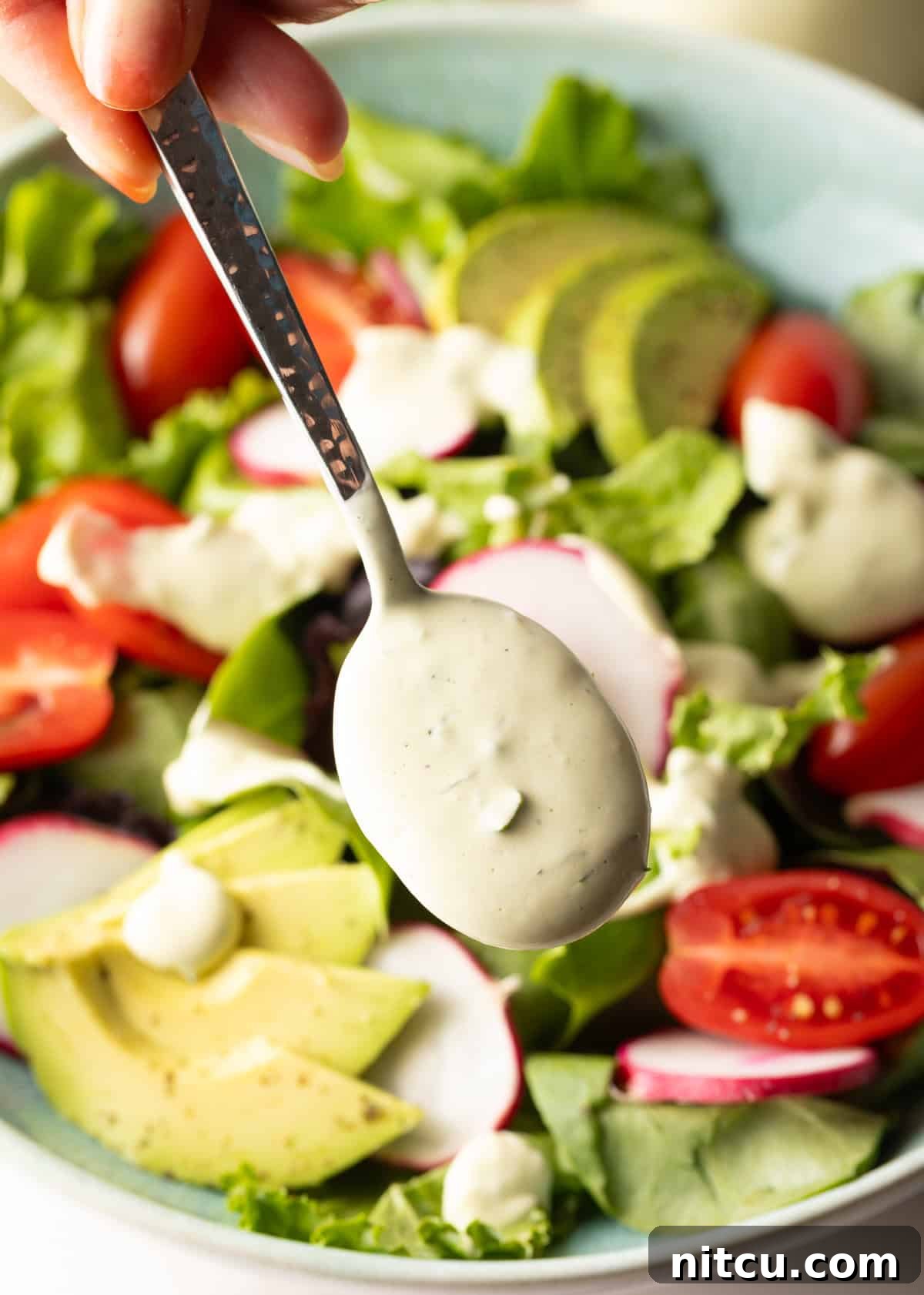 A spoon is drizzling homemade avocado lime ranch dressing over a salad with tomatoes and avocados.