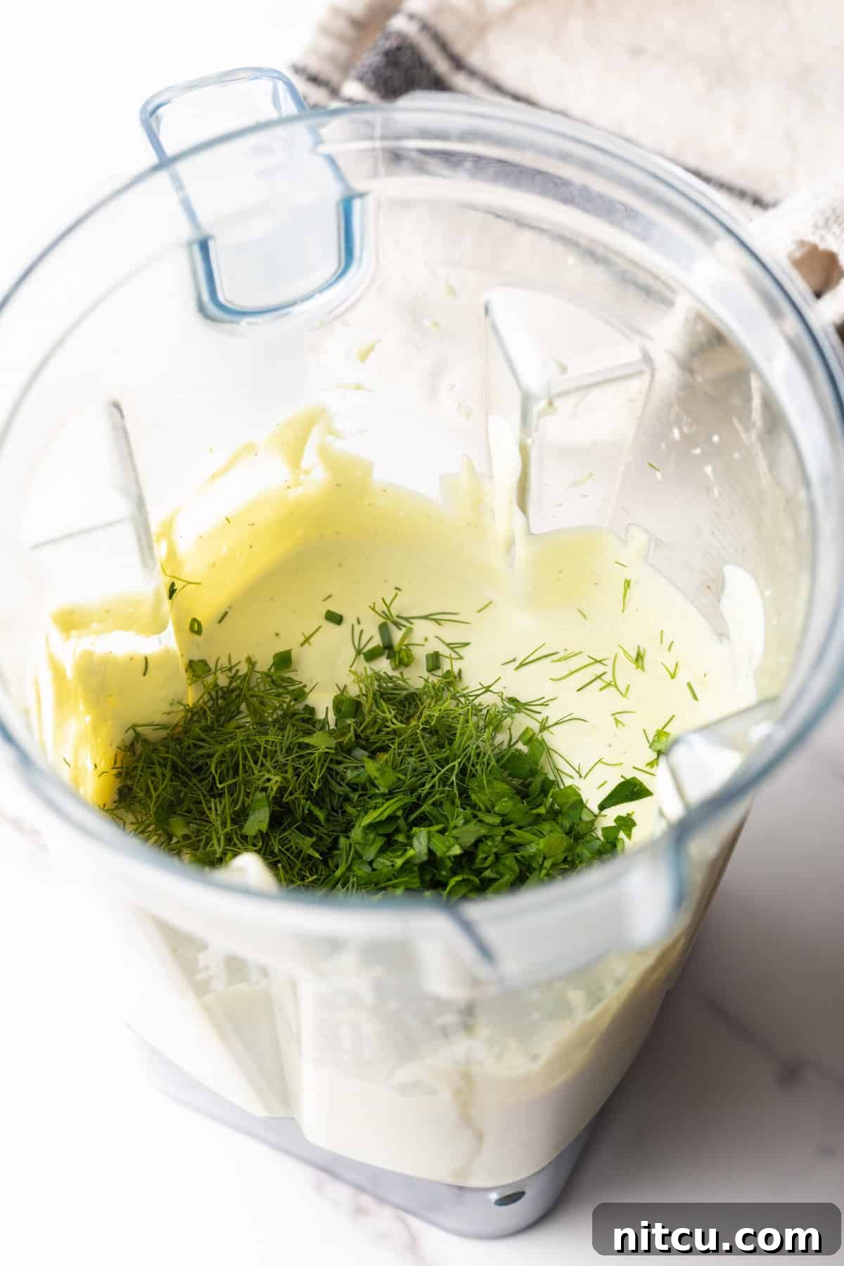 Fresh herbs are being incorporated into an avocado ranch dressing recipe with a blender.